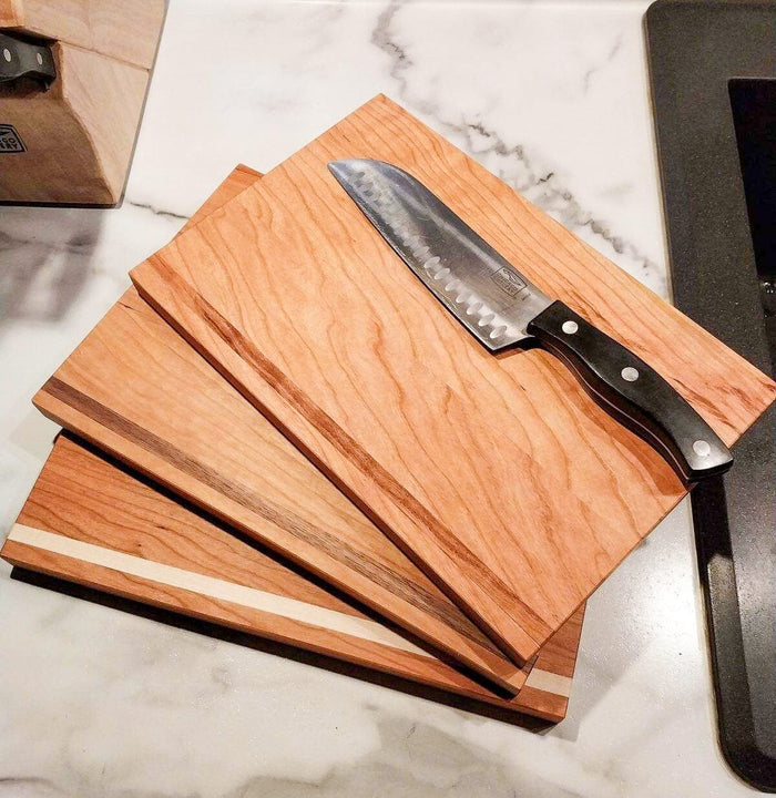 Three TY Fine Furniture Handmade Hardwood Cutting Boards are stacked on a marble countertop with a chef's knife resting on top, adding style to your kitchen. A knife block and sink can be seen in the background.