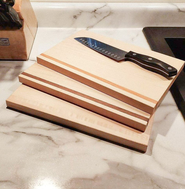 A kitchen countertop featuring the TY Fine Furniture Handmade Hardwood Cutting Board Set of three stacked boards with a black-handled chef’s knife on top. A knife block and black sink are also partially visible.