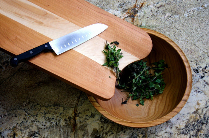 A kitchen knife sits on the TY Fine Furniture Mixed Wood Cutting Board with Integrated Bowl Scoop, beside fresh green herbs—some placed in the built-in wooden bowl on a stone countertop.