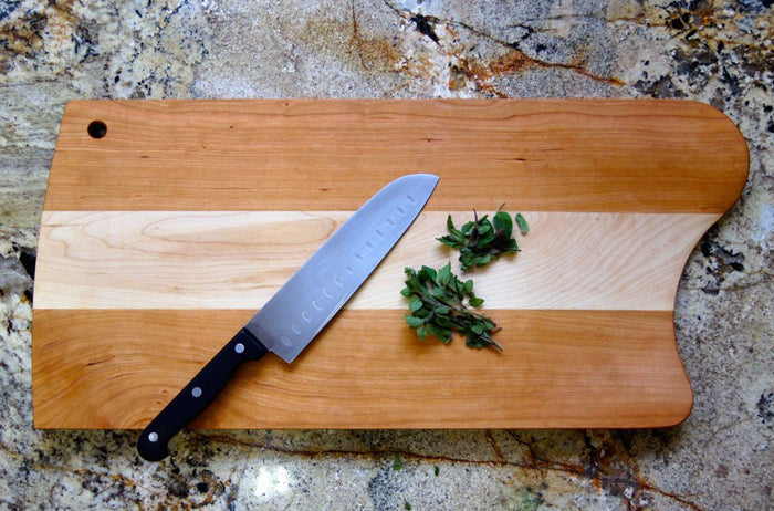 A TY Fine Furniture Mixed Wood Cutting Board with Integrated Bowl Scoop holds a chef’s knife with a black handle, beside fresh green herbs, on a marbled countertop.