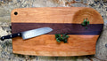 A chef’s knife and fresh green herbs sit on the TY Fine Furniture Mixed Wood Cutting Board with Integrated Bowl Serving Tray Large, featuring a dark stripe on a stone countertop.