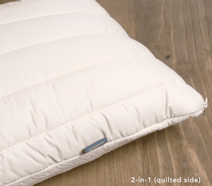The Naturepedic Organic Adjustable Latex 2 in 1 Pillow, featuring a white quilted cover and small fabric tag, rests on a wooden surface. "2-in-1 (quilted side)" text appears in the corner.