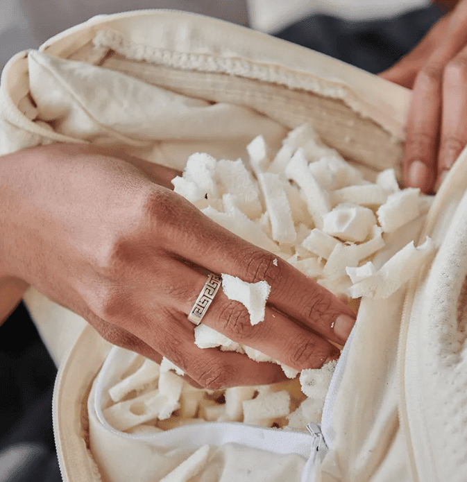 A hand wearing a silver ring reaches into a Naturepedic Organic Adjustable Latex 2 in 1 Pillow, unzipping it to reveal small, white foam pieces inside.
