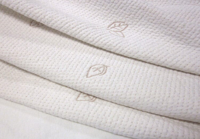 A close-up of the Organic Stretch Mattress Protector by TY Sleep, made from soft organic cotton in cream, with a subtle dotted texture and delicate light brown embroidered leaf motifs.