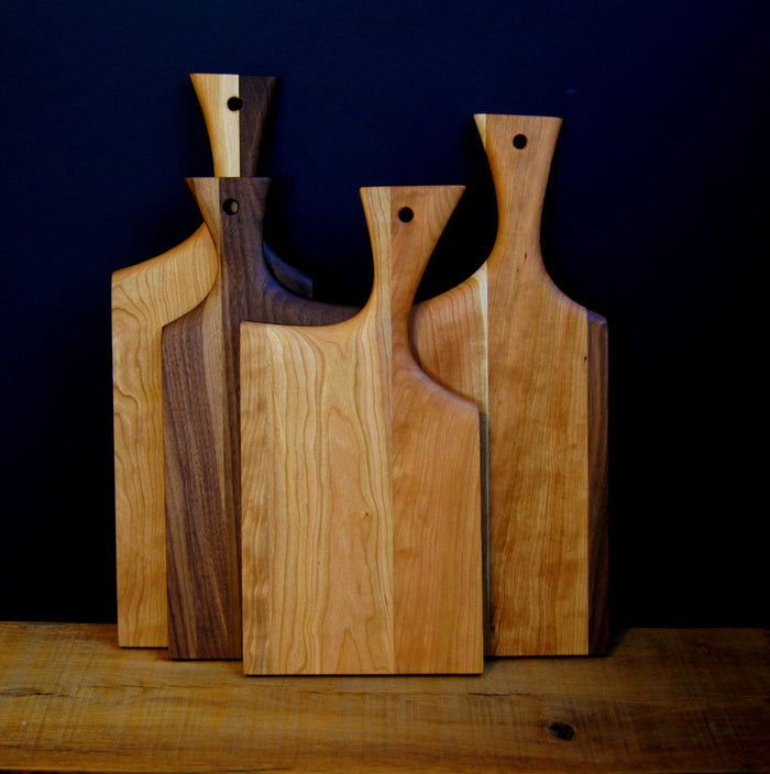 Three TY Fine Furniture Paddle Mixed Wood Cutting Boards, each with a unique handle and rounded hole, stand upright on a wooden surface. The boards showcase a range of wood tones from light to dark brown.