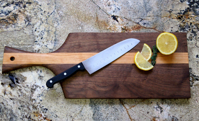 The TY Fine Furniture Paddle Mixed Wood Cutting Board, shown with a kitchen knife and two lemon halves on it, rests on a marbled countertop.