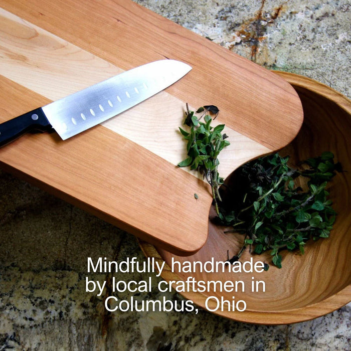 A sharp kitchen knife rests on a Solid Wood Butcher Block Cutting Board by TY Fine Furniture. Fresh herbs are chopped, some falling into a wooden bowl. Text: Mindfully handmade in Columbus, Ohio with a food-grade wood finish.